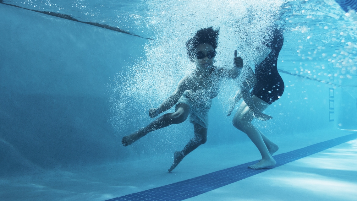 A young boy underwater in a pool giving a thumbs up next to an instructor.