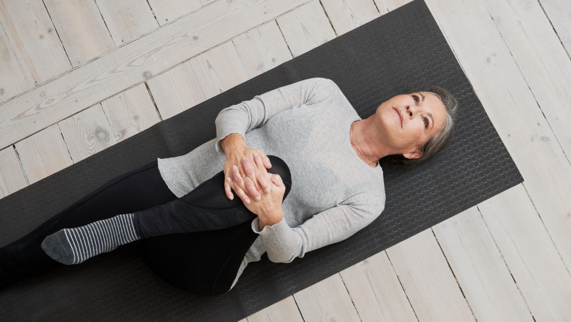 A woman lying on her back on a yoga mat with one leg stretched and one knee bent toward her chest.