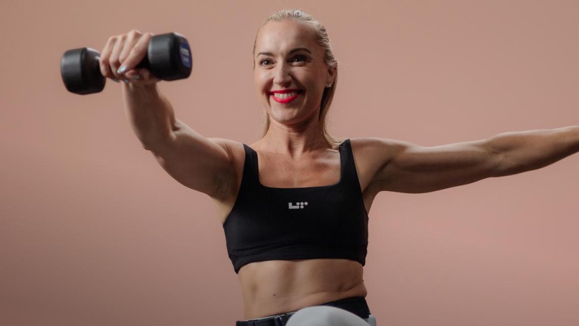 Jessie Syfko, a Life Time performer, smiling while doing an exercise move involving balancing on one leg with a dumbbell in one hand.