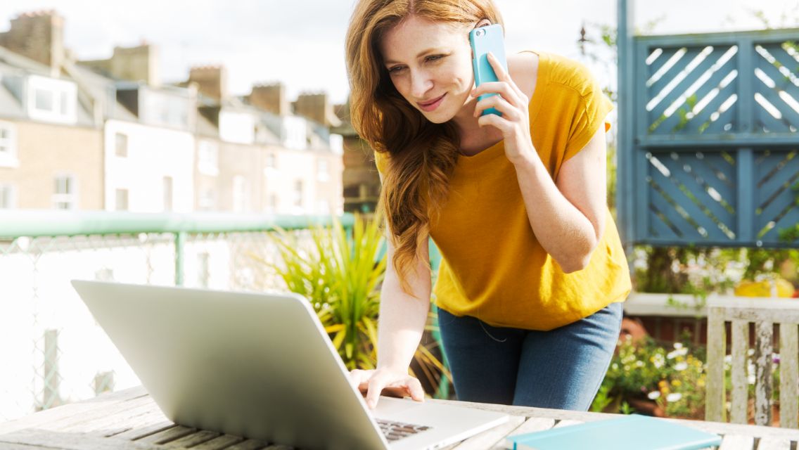 A woman working on a laptop outside while also talking on the phone.