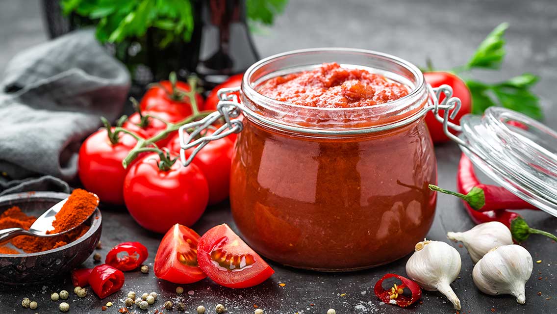 A jar of roasted tomato salsa surrounded by the produce used to make it
