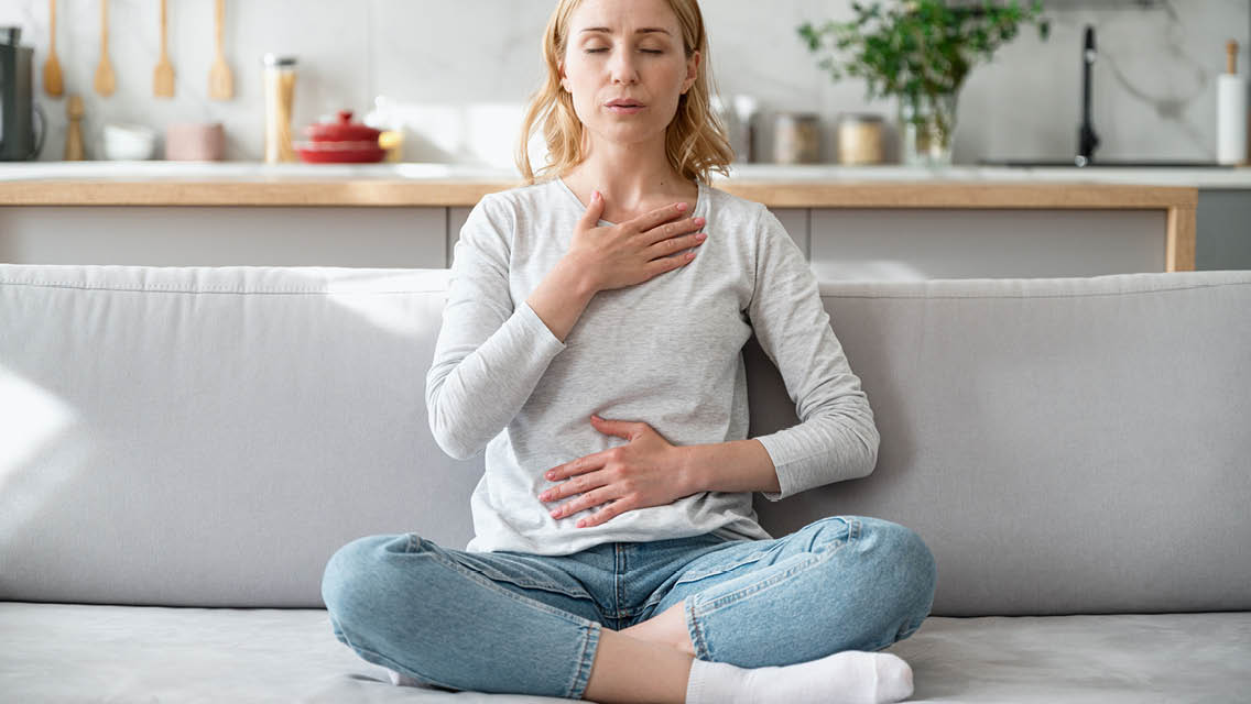 a women sits on a couch with one hand on her heart and the other on her belly performing deep breathing