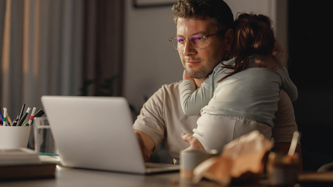 a man works on his computer with his small daughter cuddled in his arms